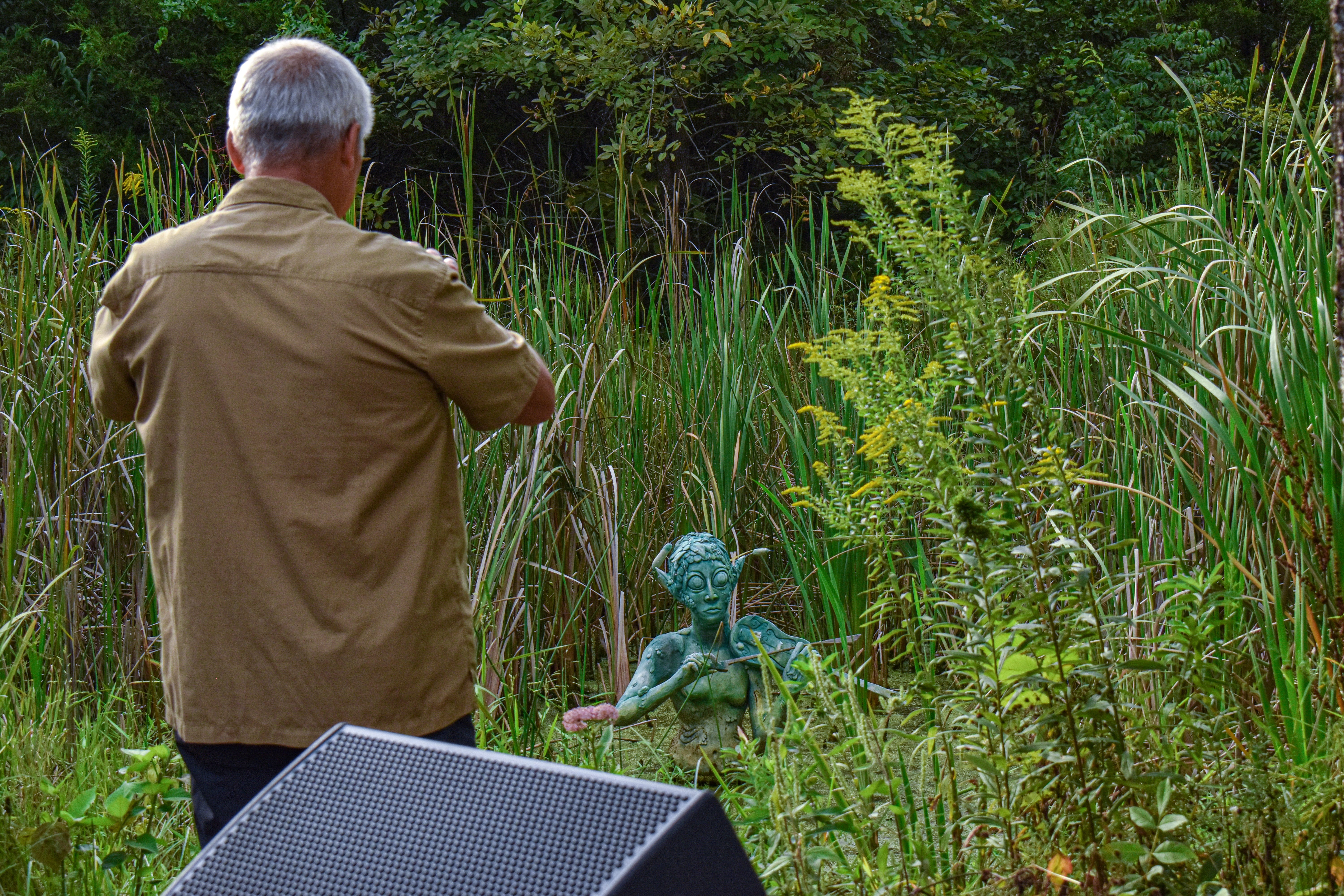 Man taking a picture of a sculpture in the pond. The sculpture is a green, somewhat fishlike, person.