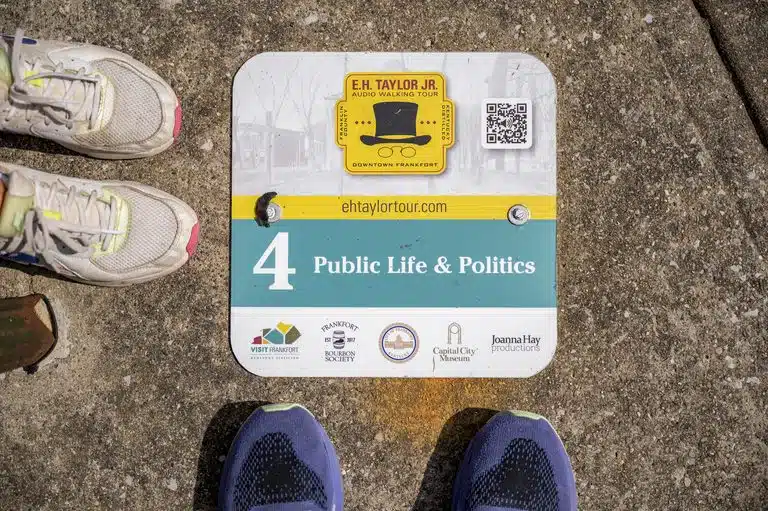 A downtown sidewalk sign for the self-guided Col. E.H. Taylor, Jr. Audio Walking Tour featuring a QR code to scan for bourbon history in Frankfort, Kentucky.