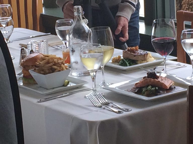 white table cloth over square table. Glasses with white wine, plates with burgers, fries, salmon dish, and thick pork chop dish