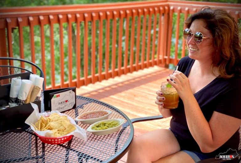 A brown-hair lady, wearing sunglasses, black shirt and bluejean shorts is enjoying an adult beverage while sitting out on the patio.  Tortilla chips, salsa and guacamole is setting on the table. 