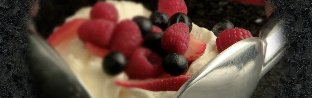 image of vanilla icecream, topped with mixed berries, between two large metal spoons on top of the granite stone.