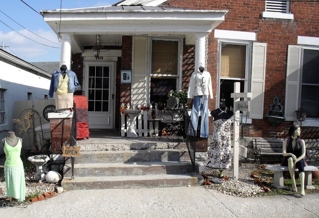 exterior shot of older brick home with items for sale displayed on the porch, steps, and sidewalk. Ms. Mary, the Mannequin, in the foreground