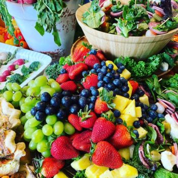 A table full of fresh fruit and vegetables. Basket is holding skewers with meat, olives, lettuce, and cheese on them. The table includes strawberries, blueberries, pineapple, grapes, bread, olives, broccoli, peppers, and more.