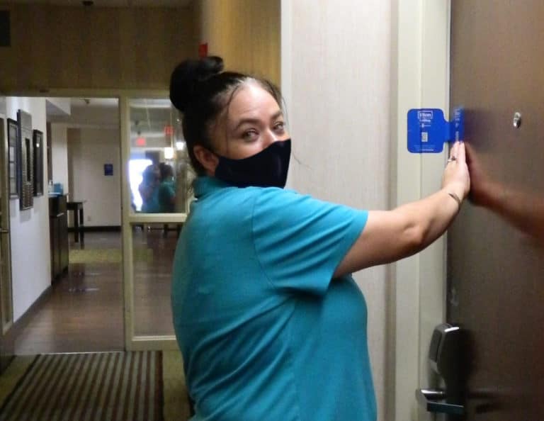 A housekeeper wearing a mask over her nose and mouth applies a seal of cleanliness on the door of a guest room at Hampton Inn Frankfort.