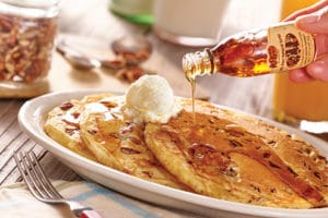 pecan pancakes on a white plate, topped with a scoop of butter, and a hand drizzling maple syrup from a bottle