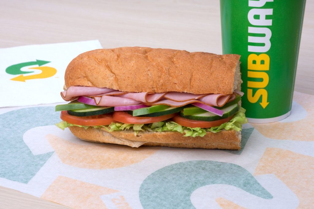 sub sandwich with ham, tomato, green peppers, and lettuce, beside a beverage cup